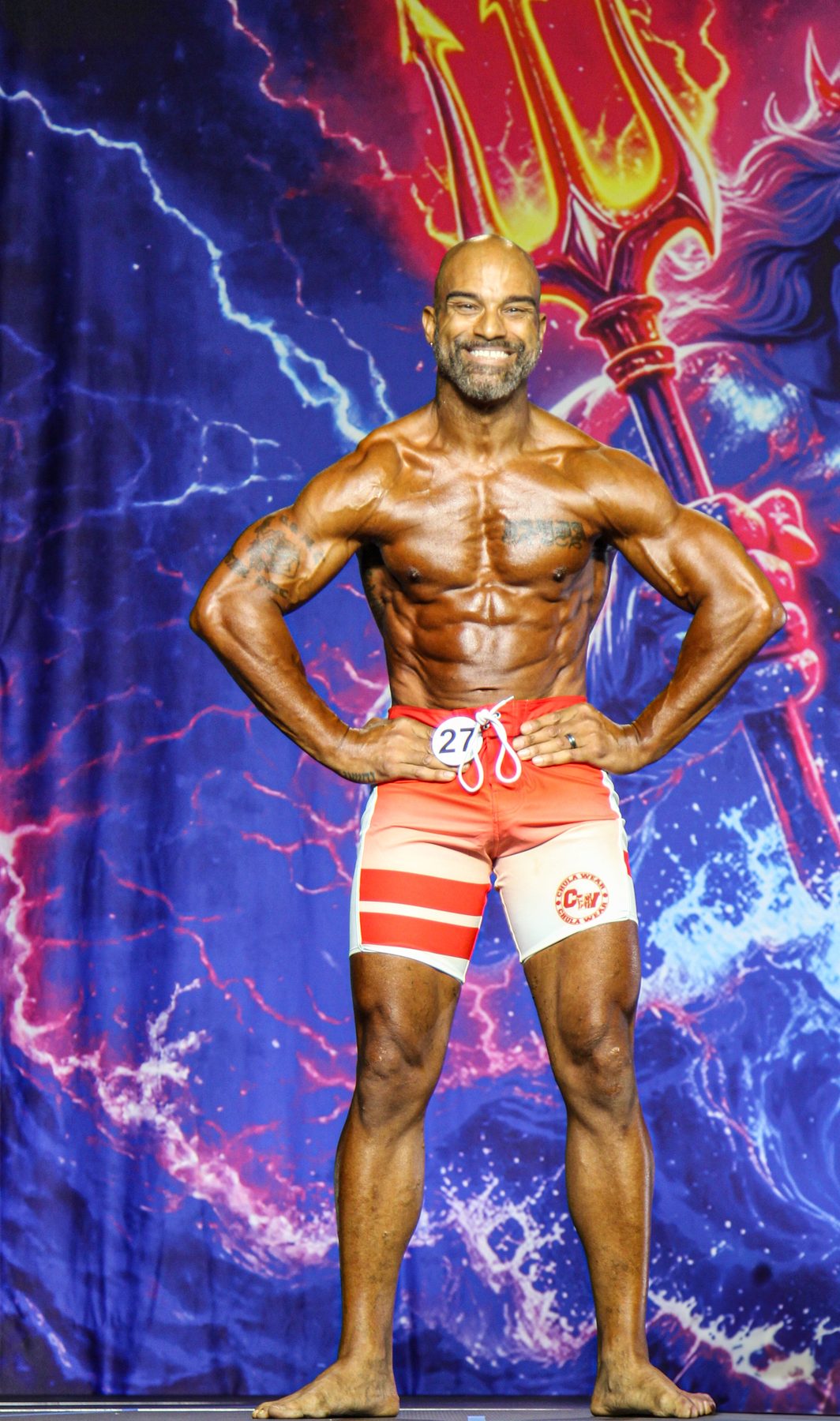 Physique competitor posing on stage under vivid backdrop lighting