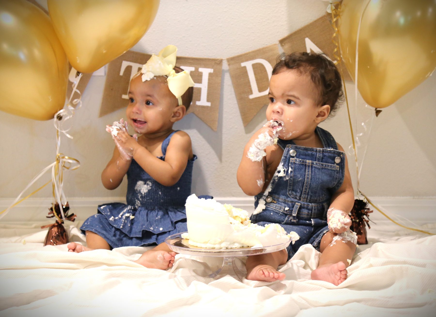Twin babies enjoying a birthday cake smash with gold balloons