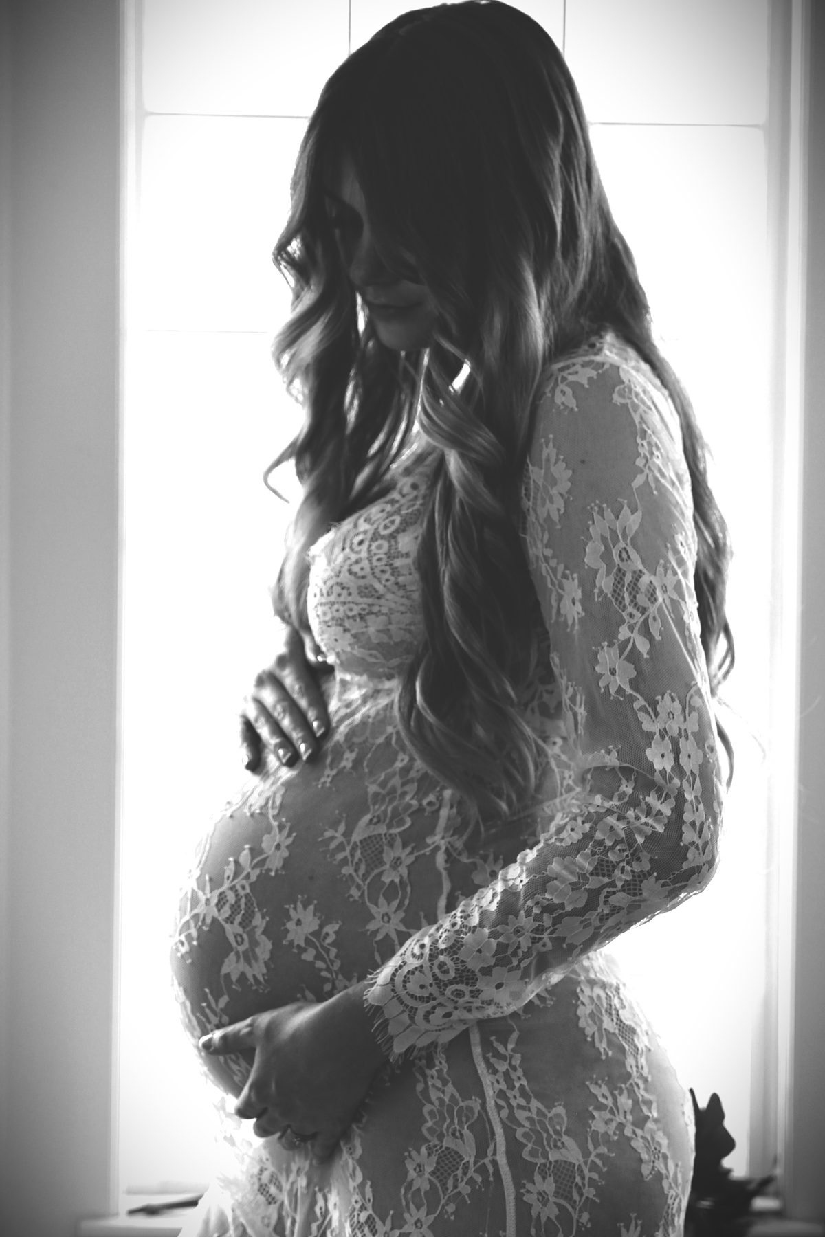 Black-and-white maternity portrait in a lace gown, silhouetted against a bright window