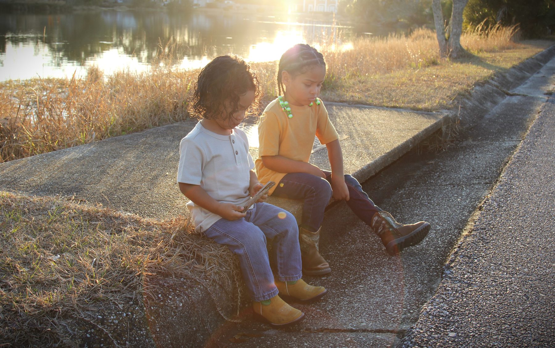 Two young siblings sitting together on a waterside curb at golden hour